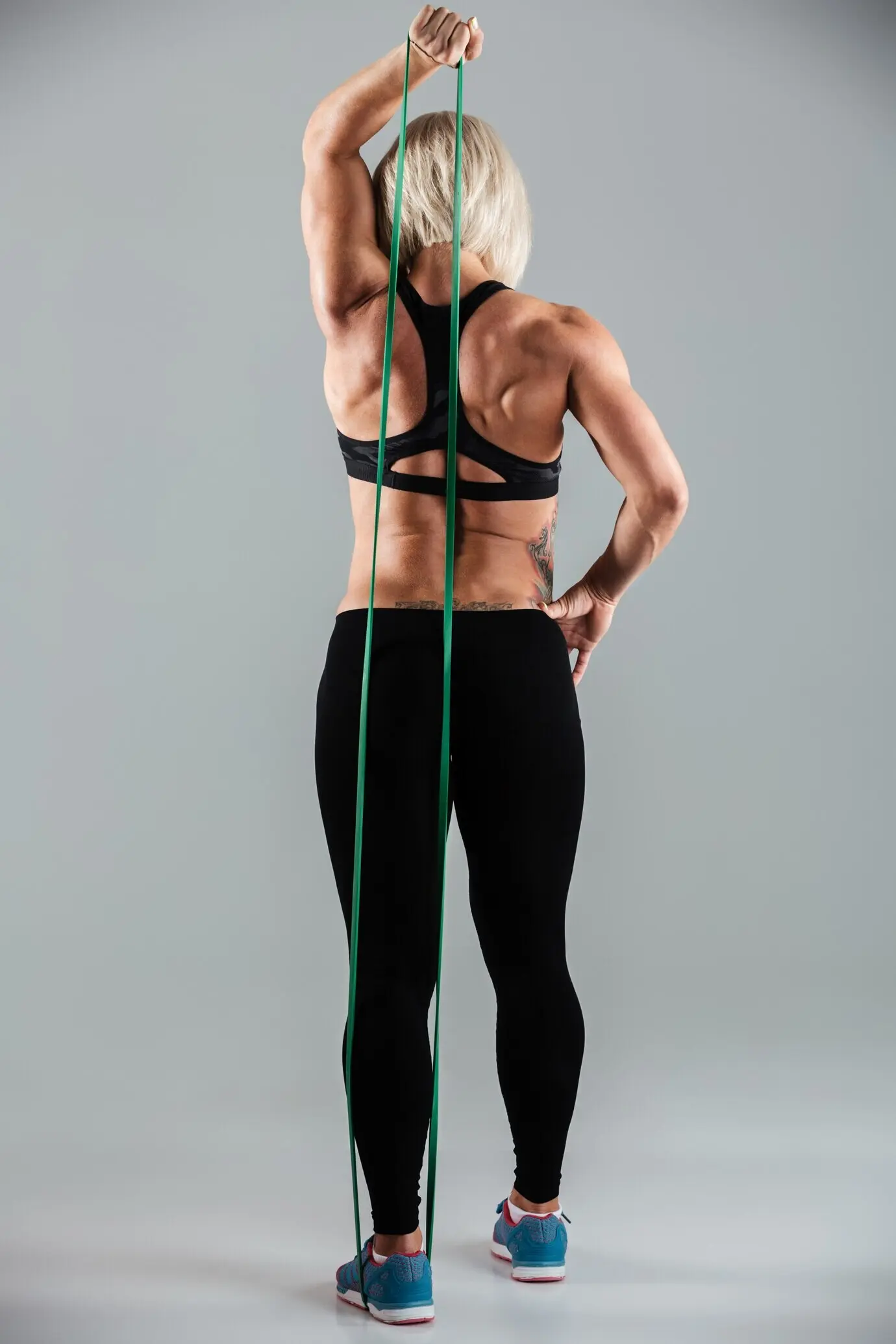 Спортсменка в спортивной одежде, снятая в полный рост, растягивает руку с помощью эластичной резинки.